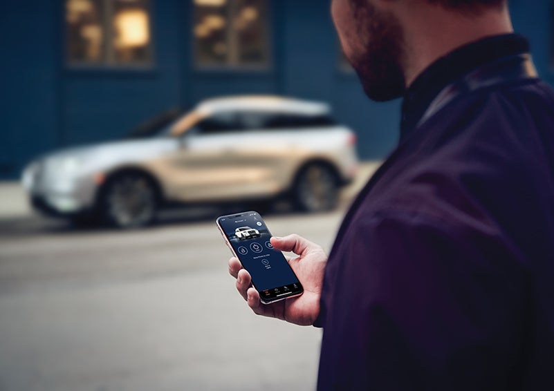 A person is shown interacting with a smartphone to connect to a Lincoln vehicle across the street. | Lincoln Cosmos Demo in Derwood MD